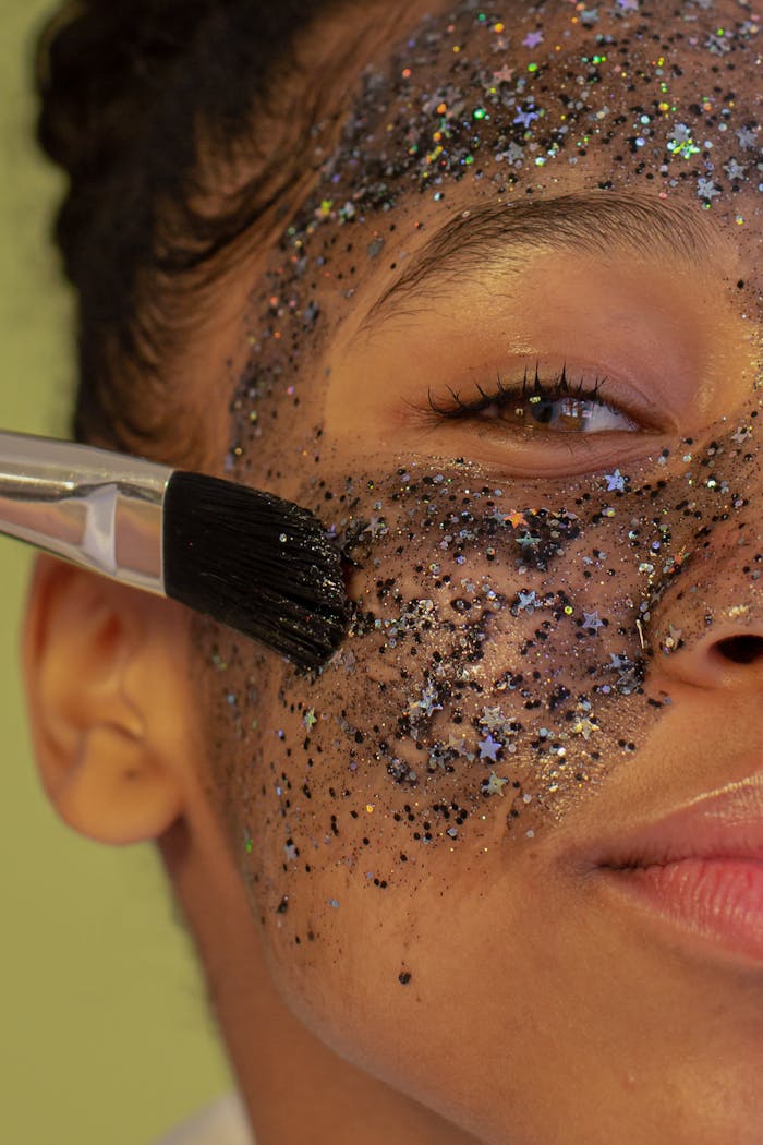 gallery-6 Crop unrecognizable young African American female with brush applying facial mask with glitters during beauty procedure and looking at camera on green background